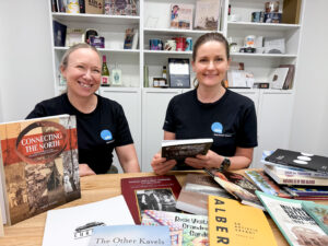 Two people seated, smiling at the camera. On a table in front of them are around 15 different books.